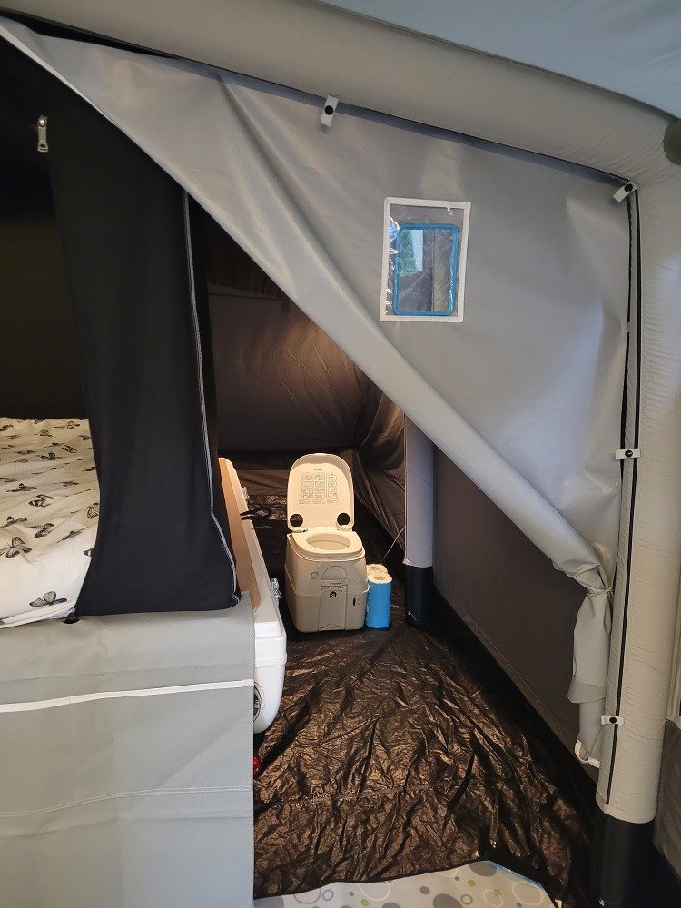 Inside a tent with a bed, portable toilet, and a small blue water container, with the tent door partly open.