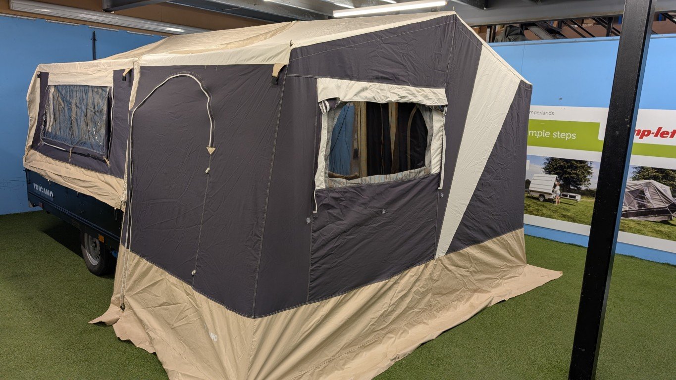A pop-up camping tent set up over a green outdoor carpeted area with a trailer attached, showing windows and a door with mesh screens.