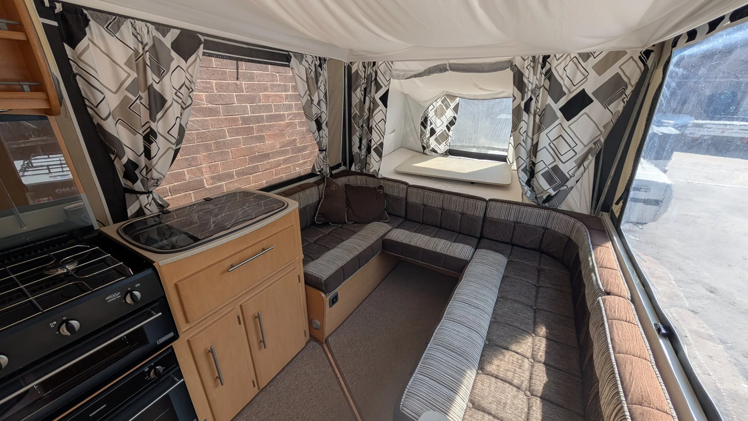 Interior of a camper trailer showing a section of a U-shaped sofa with brown and beige striped cushions, a small wooden kitchenette with a stove and a marble countertop, and windows with black and white patterned curtains.