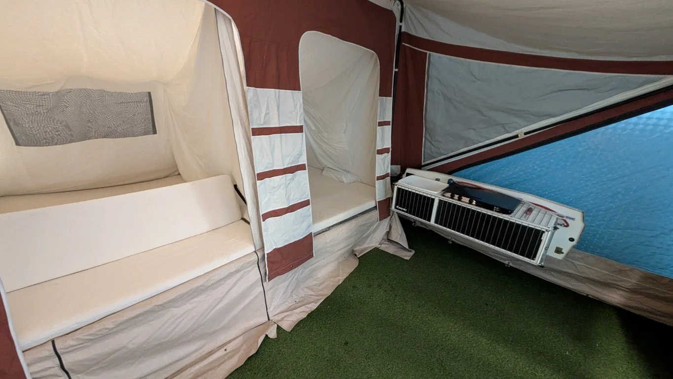 Inside a tent with a small air conditioning unit on the ground, a bed with a pillow, and a slide open window showing water outside.