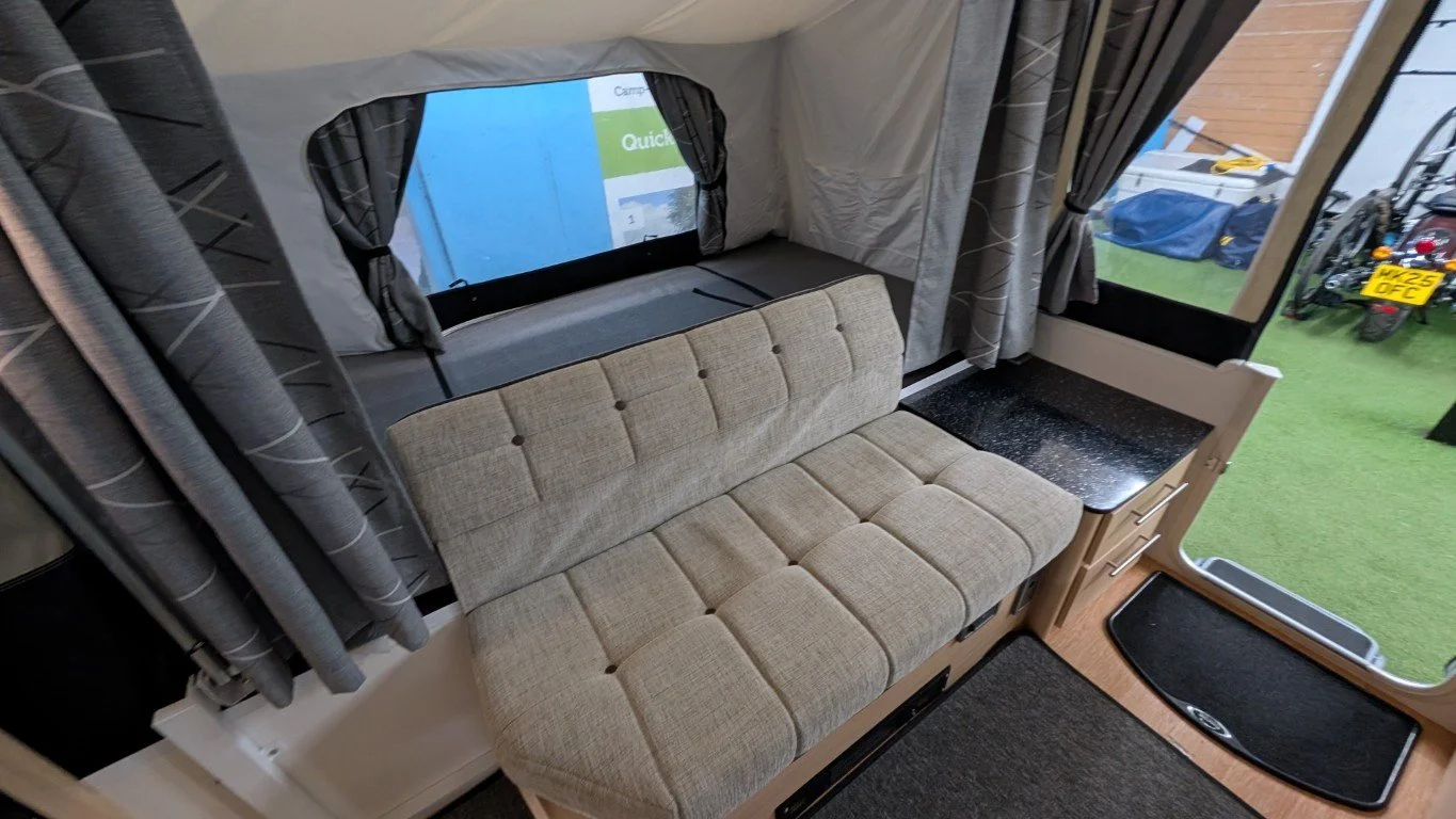 Interior of a camper or RV with a beige cushioned bench, a small countertop, and windows with curtains, with a view of the outside grass and parked bikes.