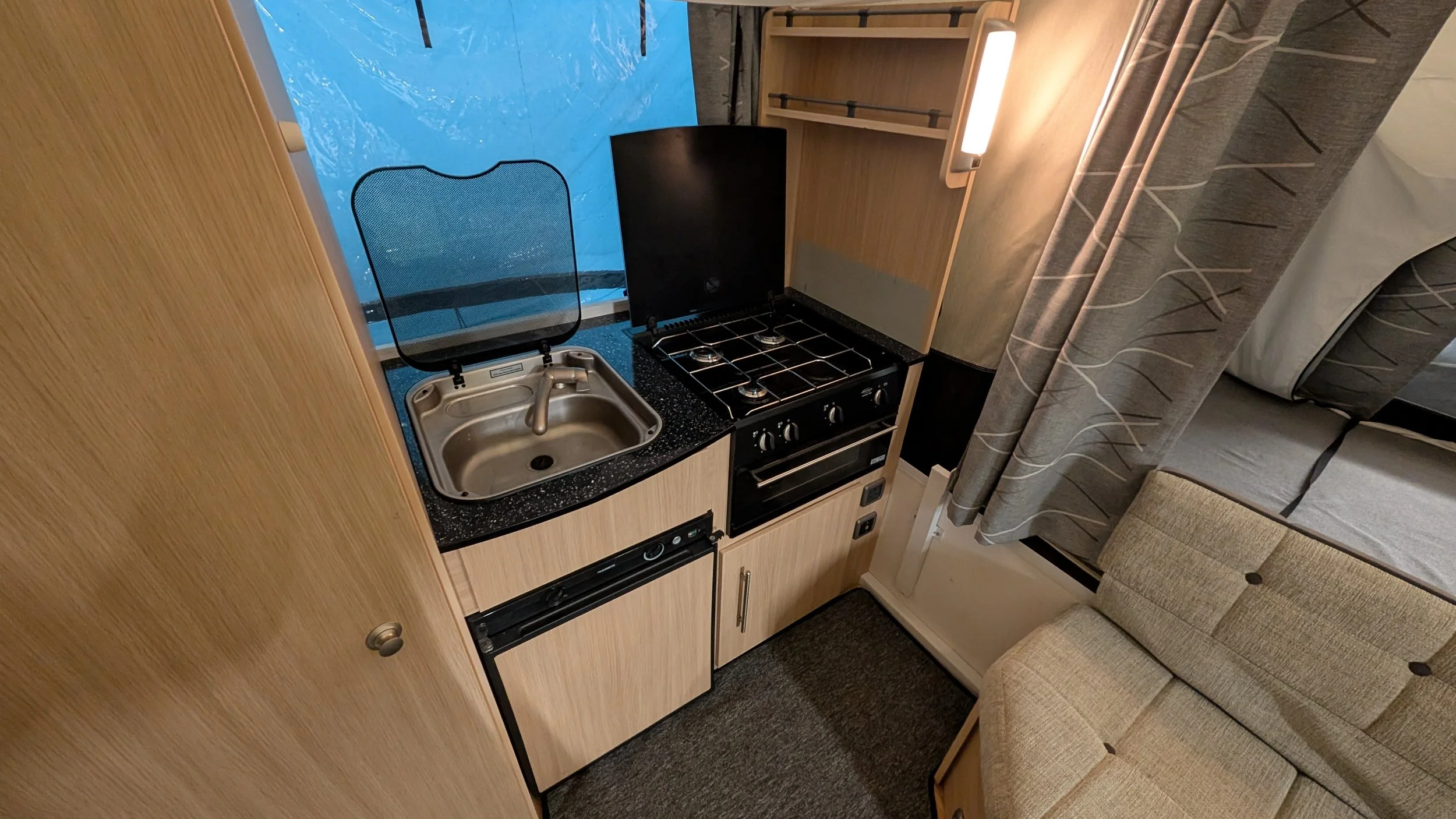 Compact kitchen area with a small sink covered by a glass lid, a black stove with four burners, a black oven beneath, and a wooden cabinet with open shelves. There's a window with a view of a boat and outdoor water. To the right, part of a beige cush