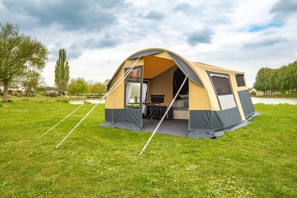 Large beige and gray Camplair tent set up on grassy area near a body of water, with trees and cloudy sky in the background.