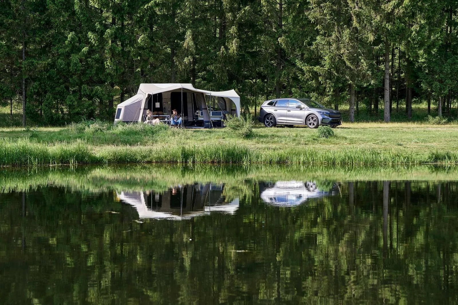 A lake with grassy banks reflects a camping scene with a tent, two people sitting under an awning, and a silver car parked nearby, surrounded by tall trees.