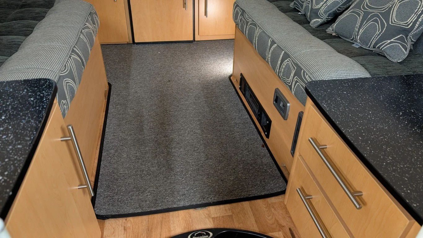Interior of a RV or camper with two cushioned bench seats facing each other, black speckled countertops, light wood cabinets, a small refrigerator, and a gray carpeted area in the middle.