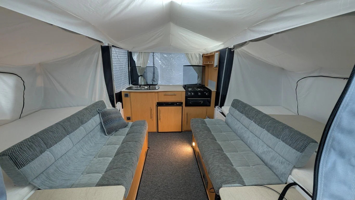 Interior of a small camper or tent with two sofas, a small kitchen area with a sink, oven, and refrigerator, and a bed at the back.
