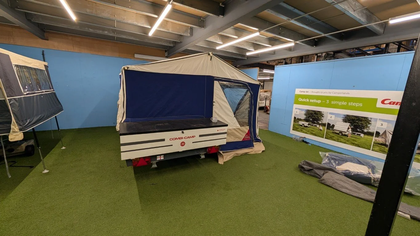 Indoor display of camping tents and accessories with a sign showing step-by-step setup instructions for a camper trailer.