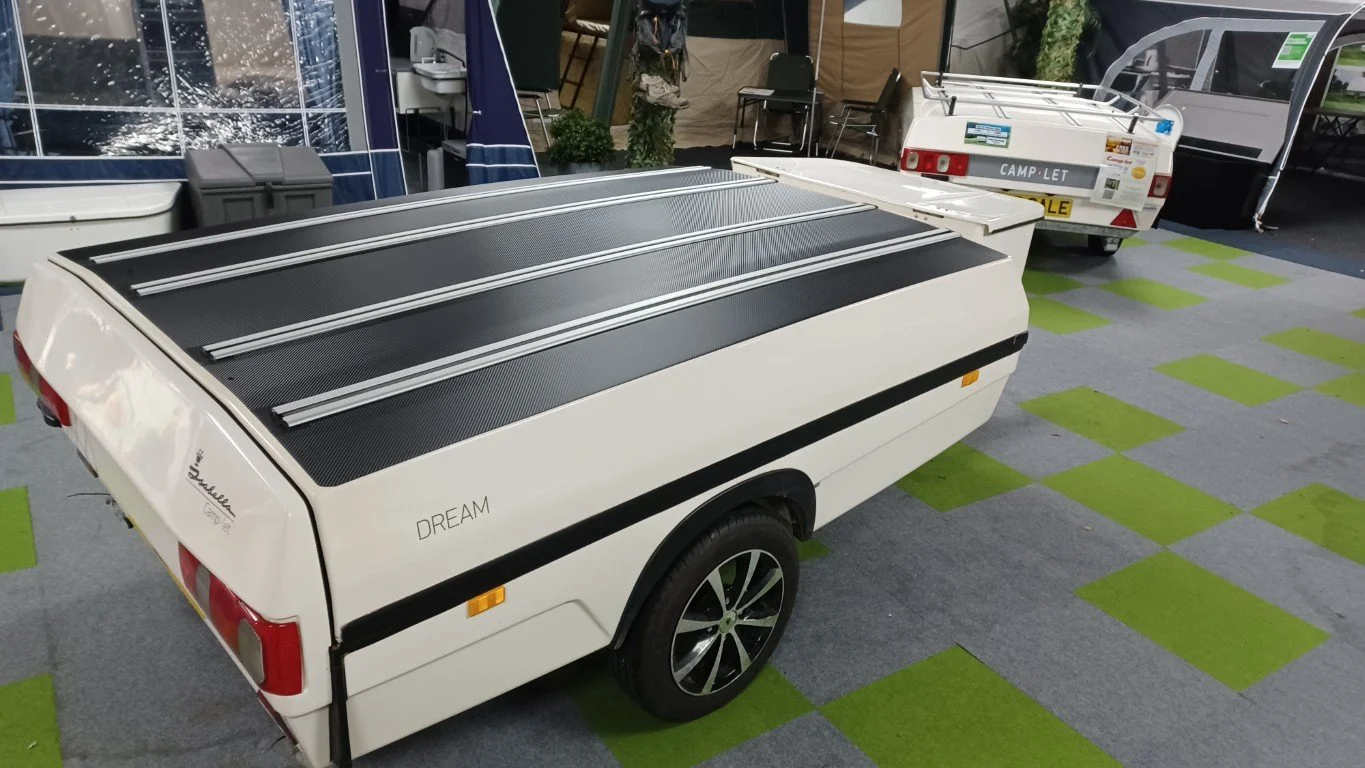 Camp-let Trailer Tent with black tops and wheel covers, displayed indoors on green and gray checkered carpet, with one labeled 'DREAM' and another labeled 'CAMP-LET'.
