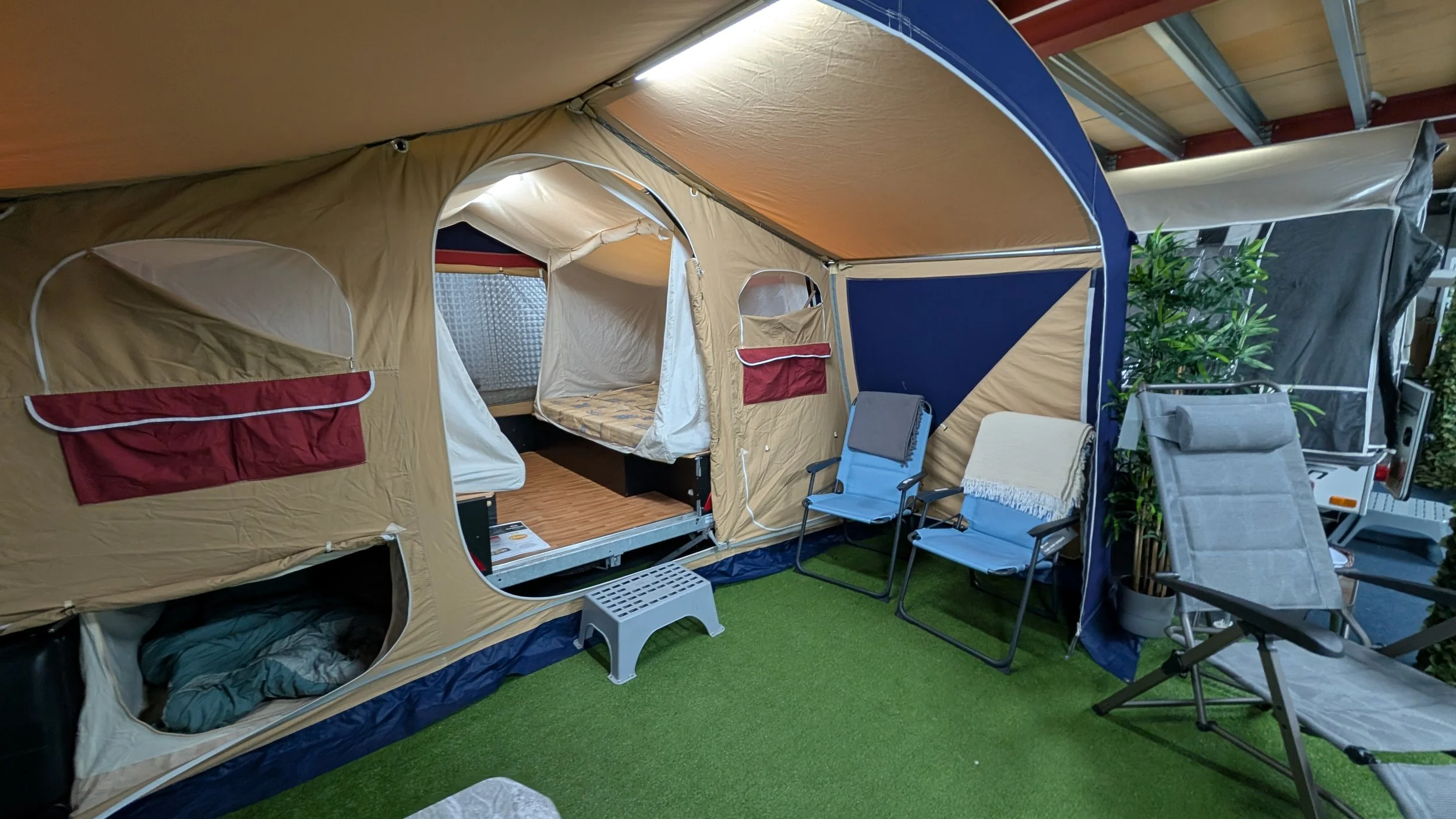 Indoor display of a large beige and blue camping tent with multiple windows and a sleeping area. Surrounding the tent are outdoor chairs, a small step stool, and artificial grass flooring, with some potted plants and additional camping equipment near
