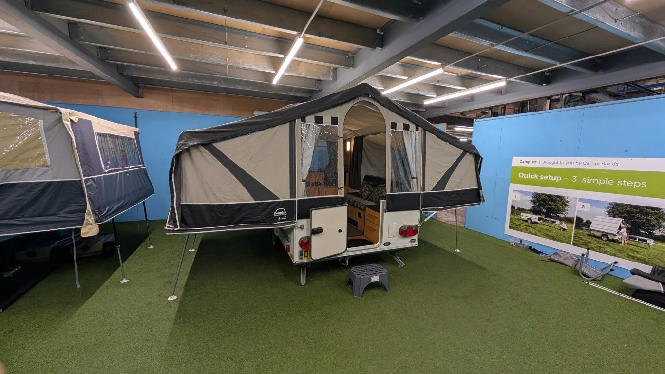 A compact camper trailer with extended tent-like sidewalls, set up indoors on artificial grass. There is a small step stool at the back and a poster on the right side explaining a quick setup process. The trailer has an open door revealing a wooden i