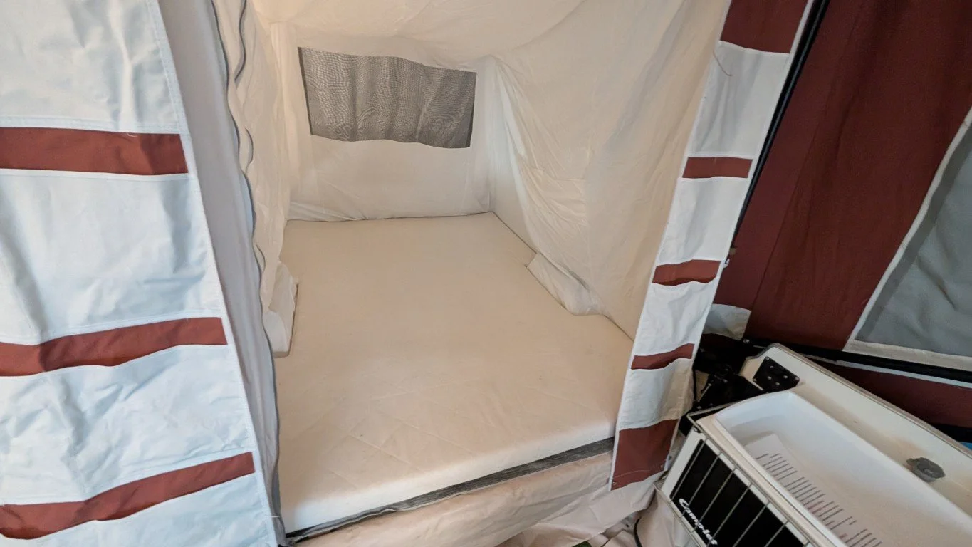 A bed inside a temporary shelter with striped fabric walls, a window, and a small heater or cooler nearby.