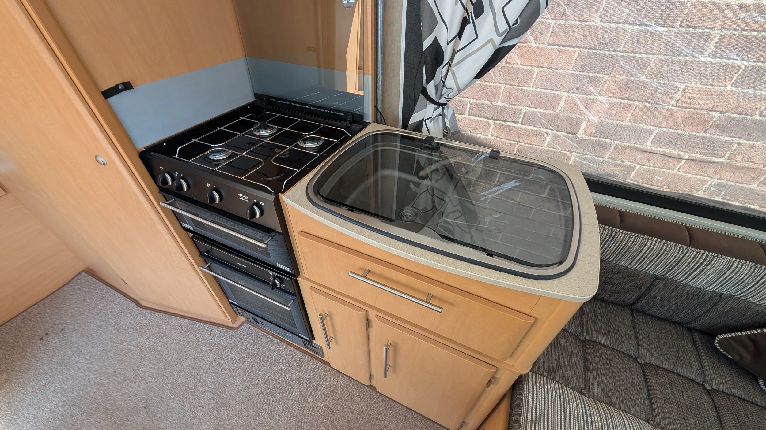 Compact kitchen area with a small gas stove, oven, and a countertop with a sink cover, next to a window overlooking a brick wall, in a cozy living space.