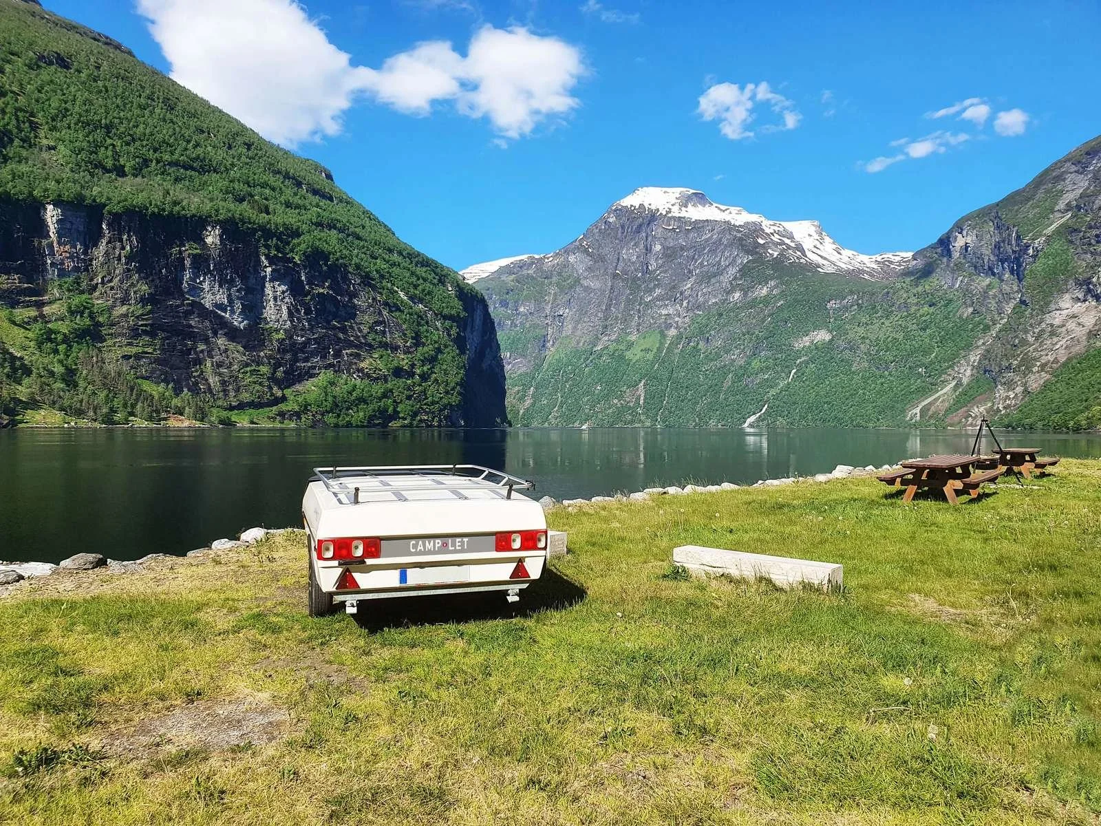 A scenic lakeside with a Camp-let trailer, picnic tables, and mountains in the background under a partly cloudy sky.