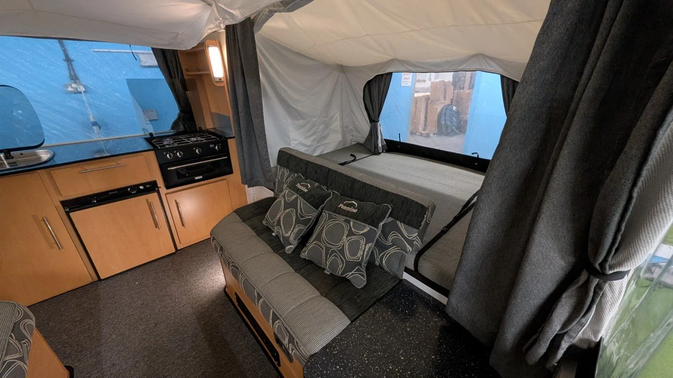 Inside a camper trailer showing a small kitchenette with a sink, stove, and mini refrigerator, a sofa with patterned cushions, and a large window with curtains.