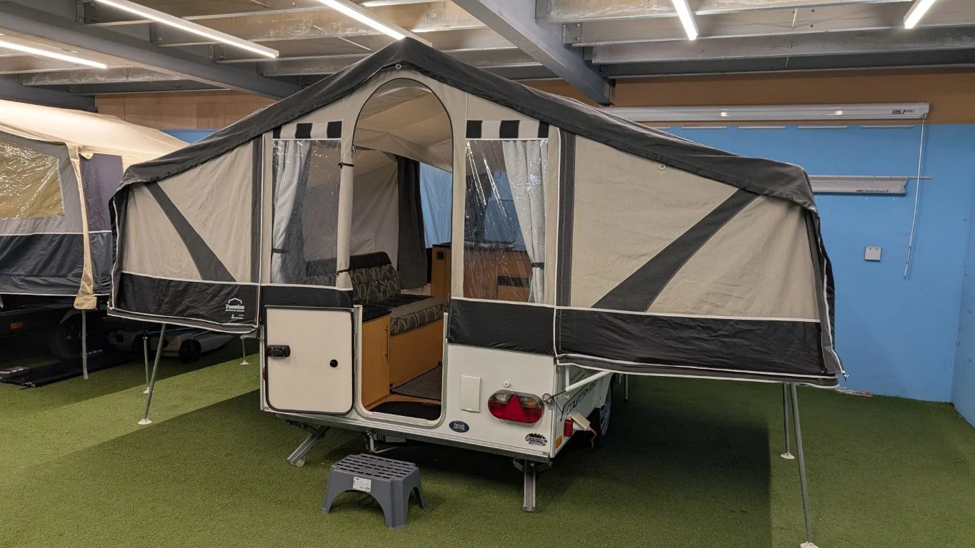 A small pop-up camper trailer with an extended tent attachment, set up indoors on artificial turf.
