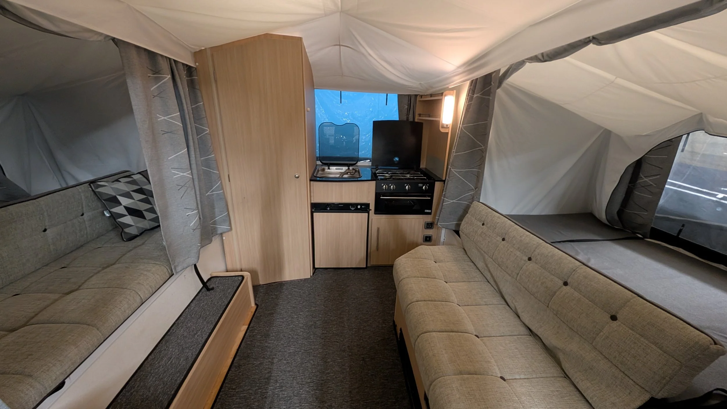Interior of a compact camper trailer with beige couches on both sides and a small kitchen area with a stove and sink in the center, with large windows covered by curtains.