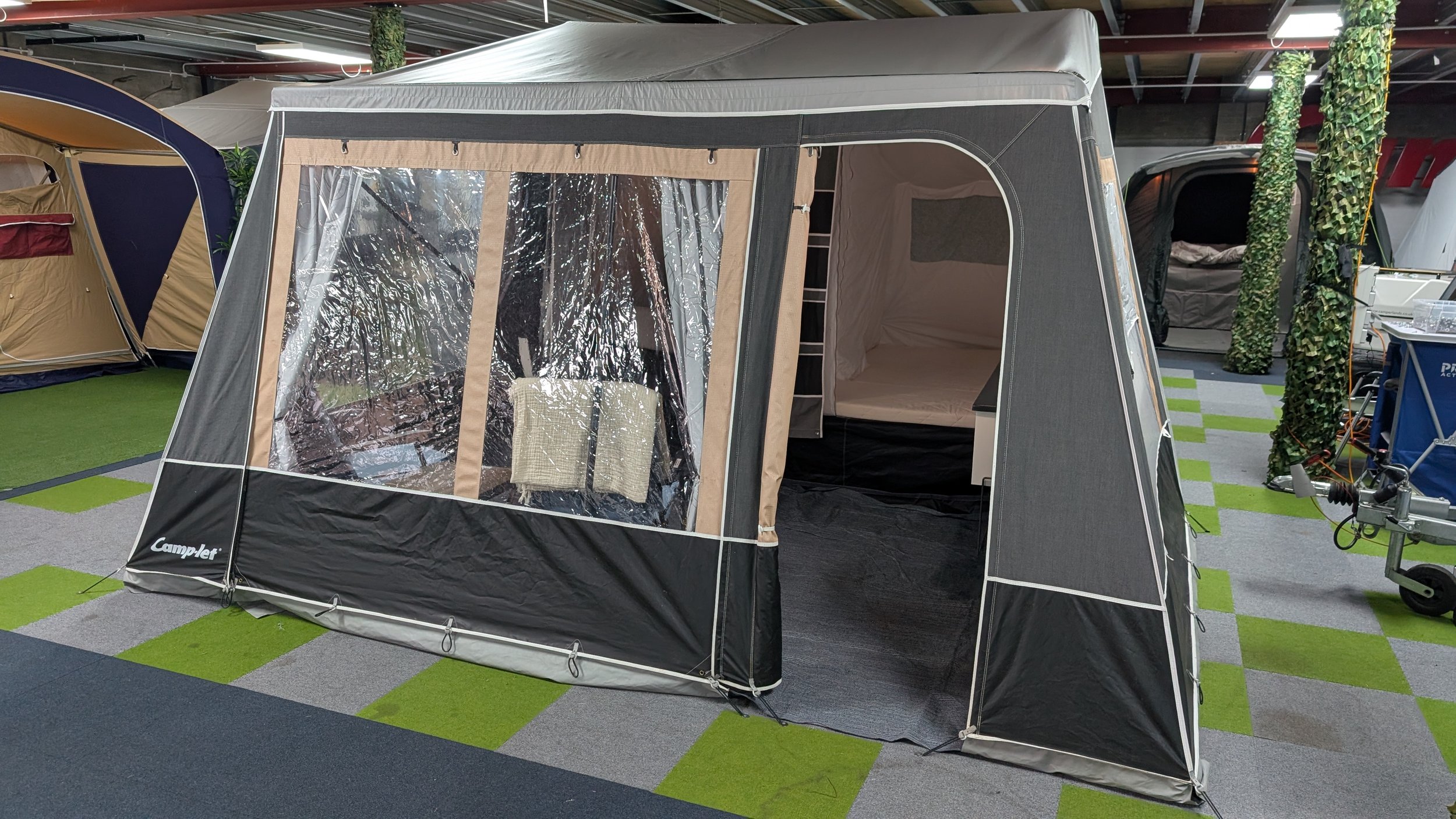 A black and beige portable camping tent with a clear plastic window on the front, set up indoors on a patterned green and gray carpet, with other tents and camping supplies visible in the background.