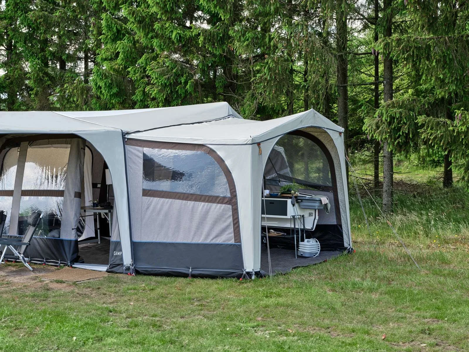 A large Camp-let tent set up in a grassy area near pine trees, with a side partially open showing outdoor dining and cooking equipment inside.