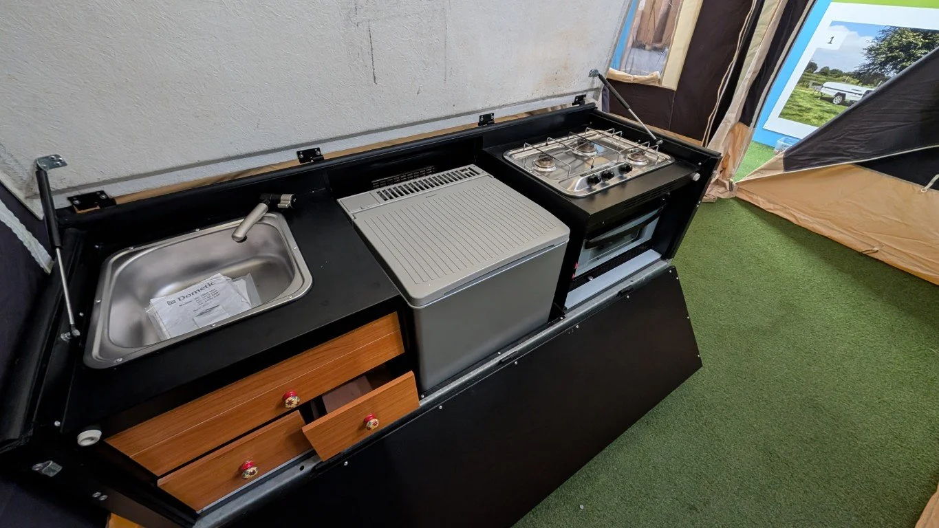 Miniature kitchen setup with a sink, a small refrigerator, a stove with burners, and cabinets, set on a green carpet indoors.