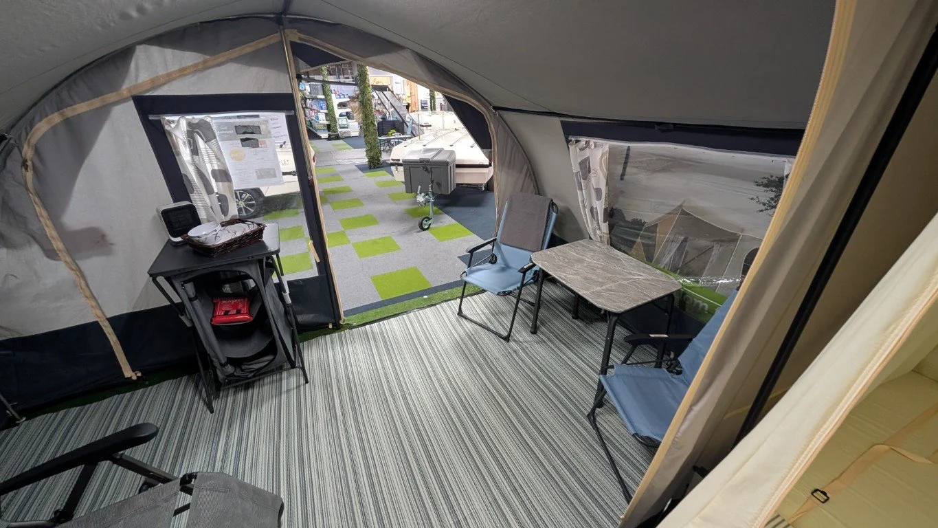 Inside a camping tent with chairs, a small table, a folding table with a basket and electronic device, and a view of the outside camping area with other tents, a trash bin, and trailers.