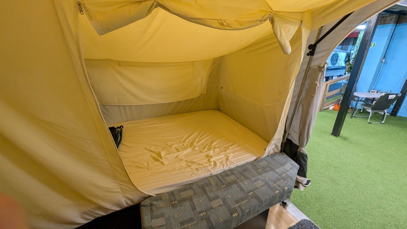 Interior of a small camping tent with a bed, and an outdoor area with green artificial grass, chairs, and a blue building in the background.