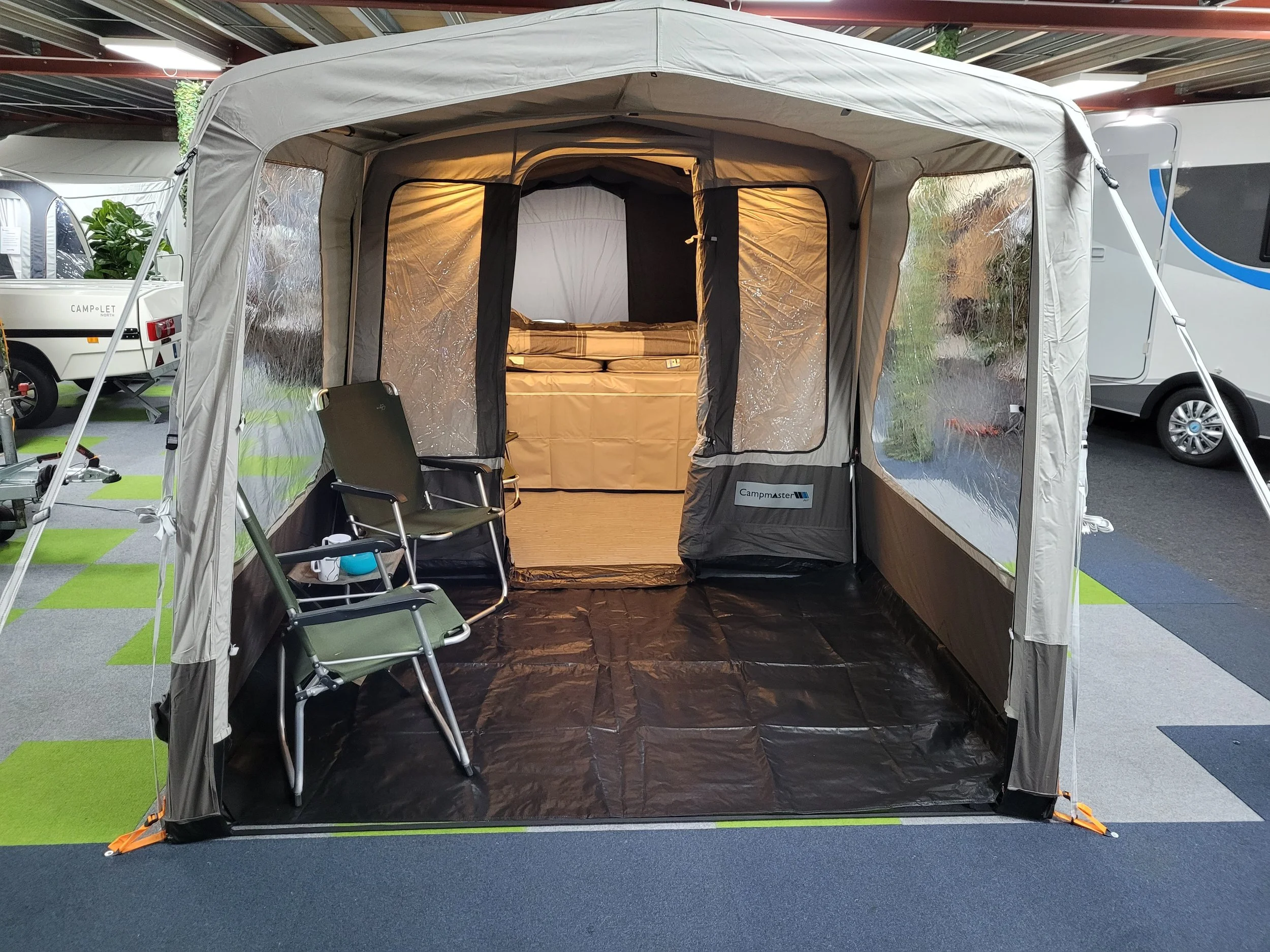 A Campamster Air tent with a black floor, two camping chairs, and a fold-up bed inside. The tent is set up in a showroom or store, with other trailers and RVs visible nearby.