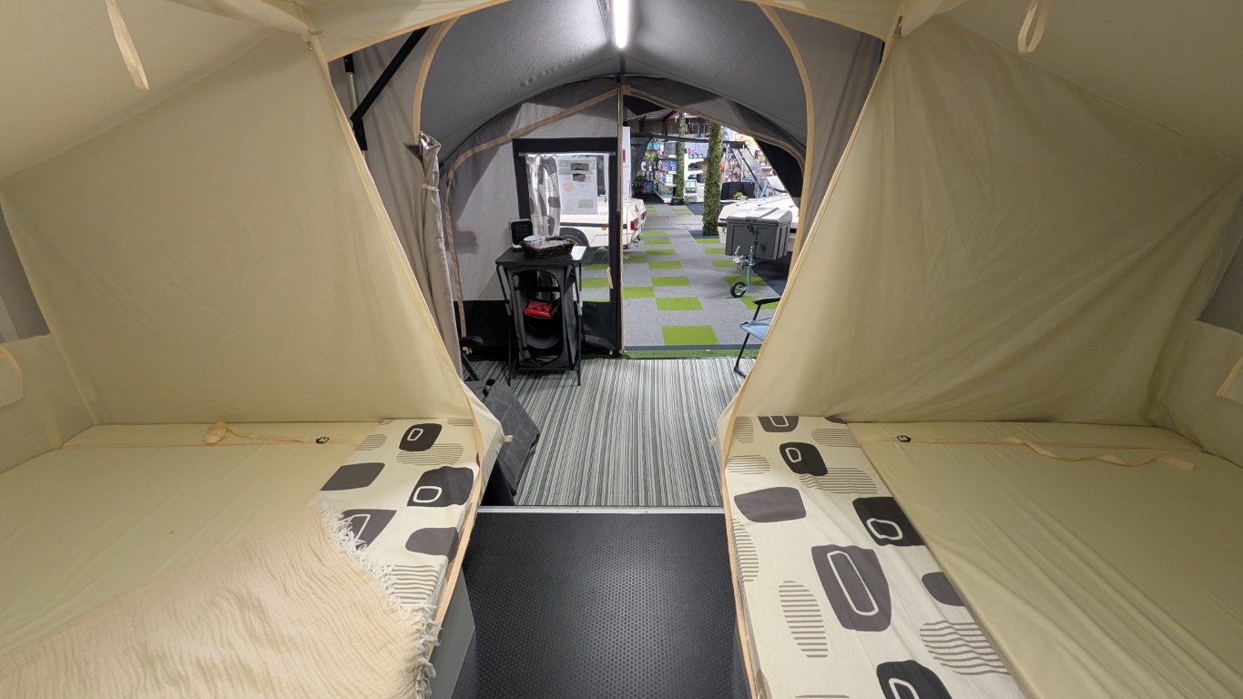 Interior of a camping tent with dual beds, a floor with striped and black textured mats, and an open flap leading to a camping site with chairs, tables, and parked vehicles outside.