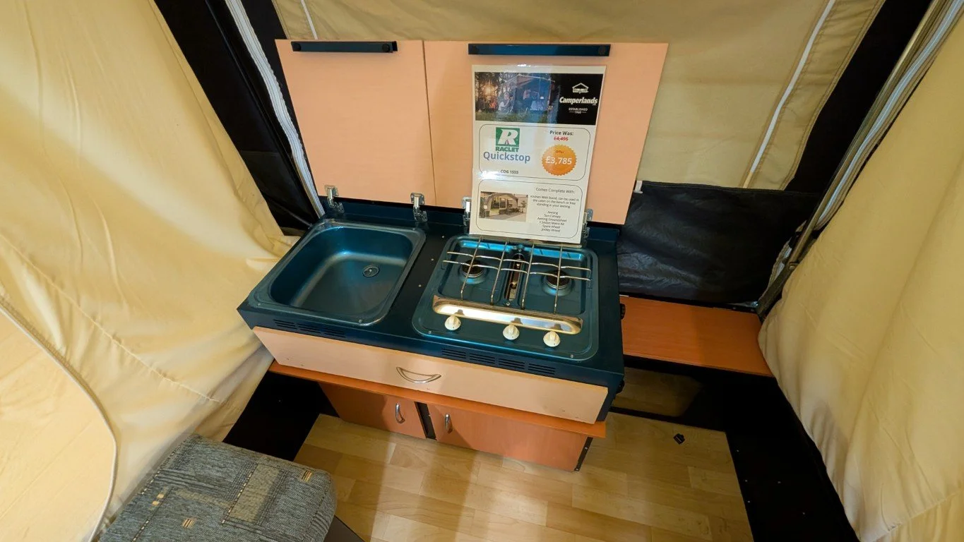 Small kitchen setup inside a camper or trailer with a sink and two-burner stove, wooden cabinetry, and seating area with yellow cushions.