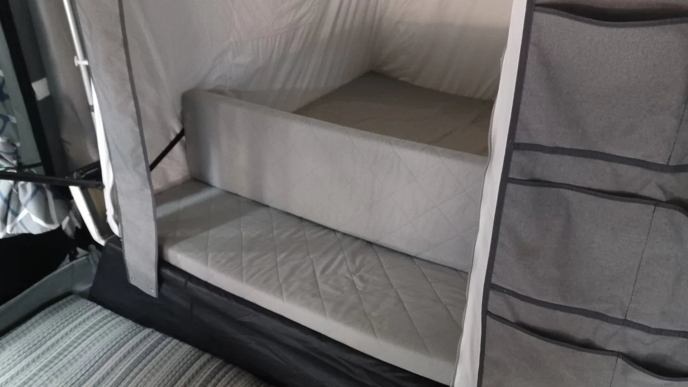 A fold-out bed inside a Camp-let Trailer Tent with a quilted mattress and a foldable cushion or backrest.