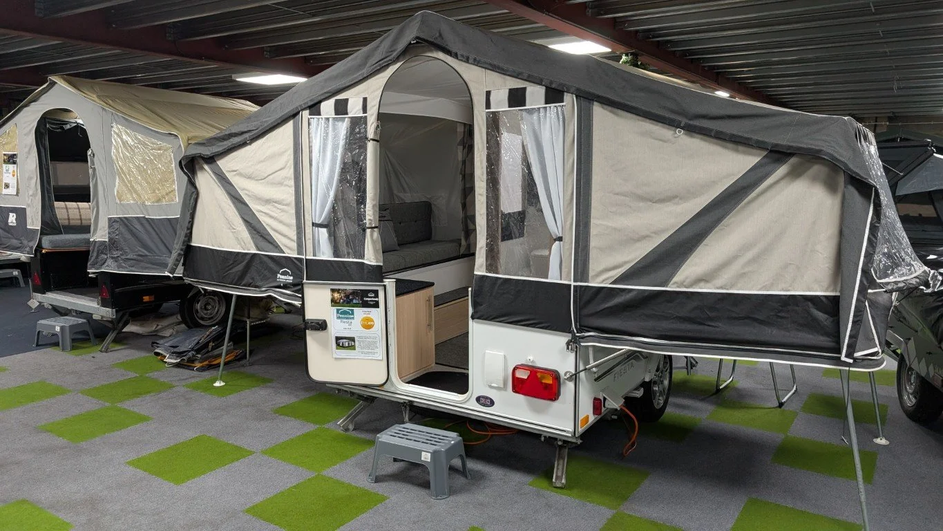 A travel trailer parked next to a brick wall, with a black and gray pop-up roof tent extended on top.
