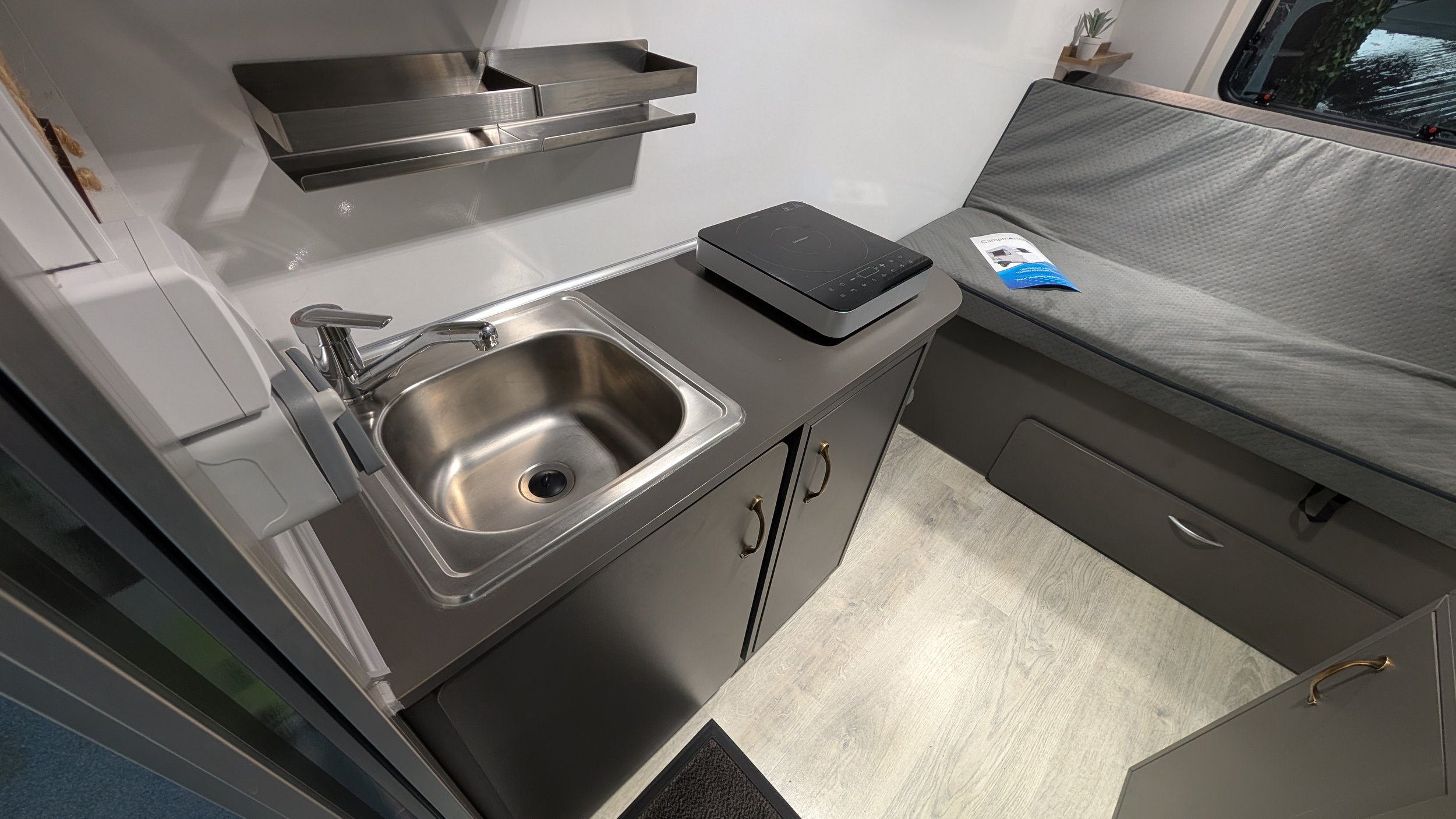 Small kitchen area with a stainless steel sink, a portable induction cooktop, and a gray upholstered bench with a cushion, located inside a compact living space, possibly a camper or tiny home.