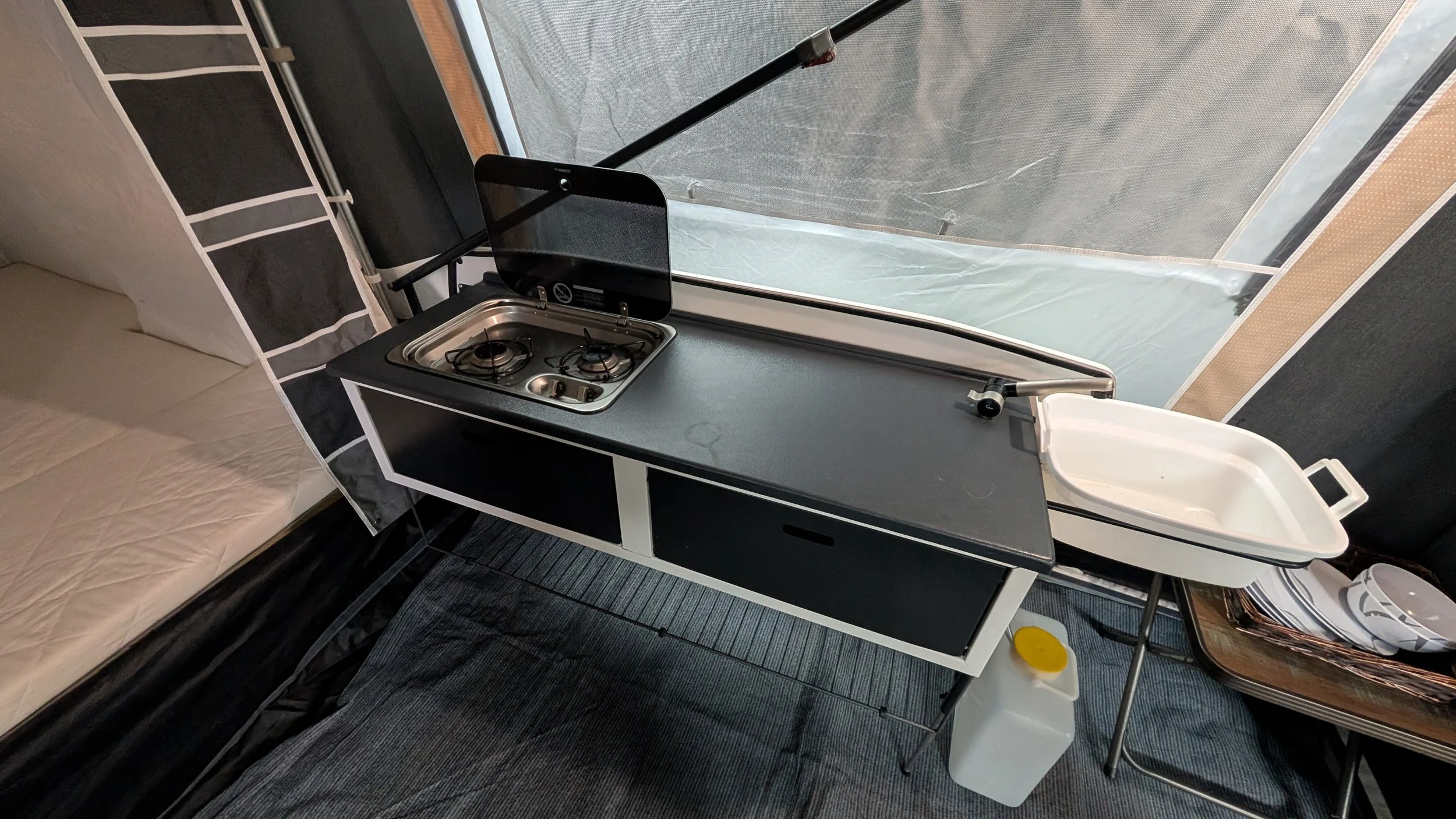 Interior of a camper van with a black countertop containing a small two-burner stove, a sink, and a glass cover over the stove. There is a reclining chair or bed with a quilted beige cover on the left and a window with a screen on the right.