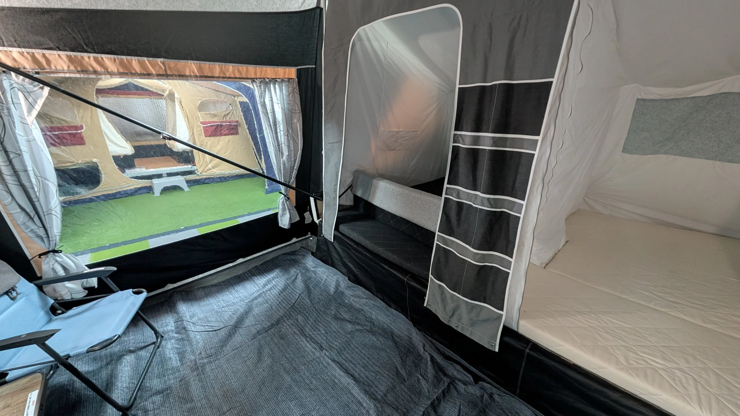 Interior of a small tent with a foldable chair, a mattress, and a bunk bed with striped sheets, and a window showing outdoor grass and building.