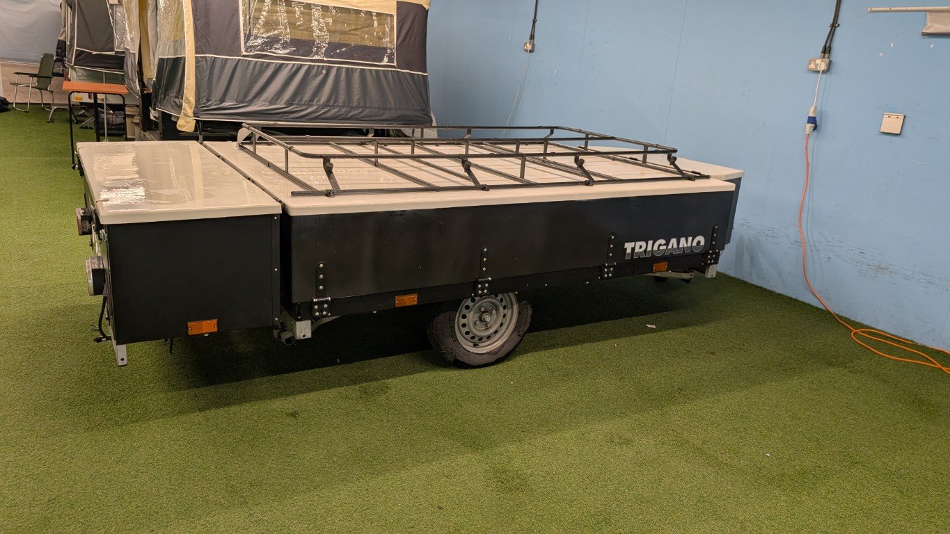 A portable black and white flatbed trailer with a metal rail on top, located in an indoor setting with green carpeted flooring and a light blue wall. The trailer has the word "TRIGANO" on its side.