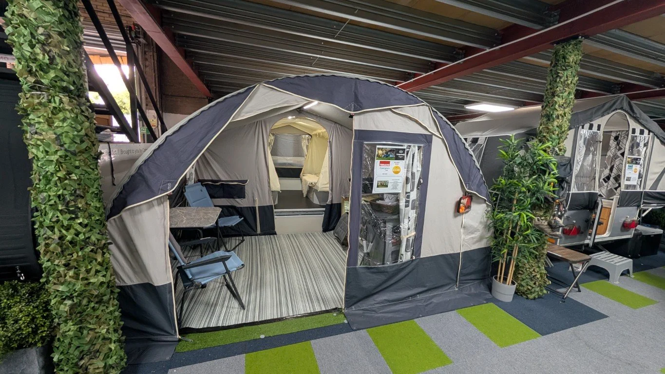 A camping tent set up indoors with an entrance, small tables, chairs, a potted plant, and display boards inside.