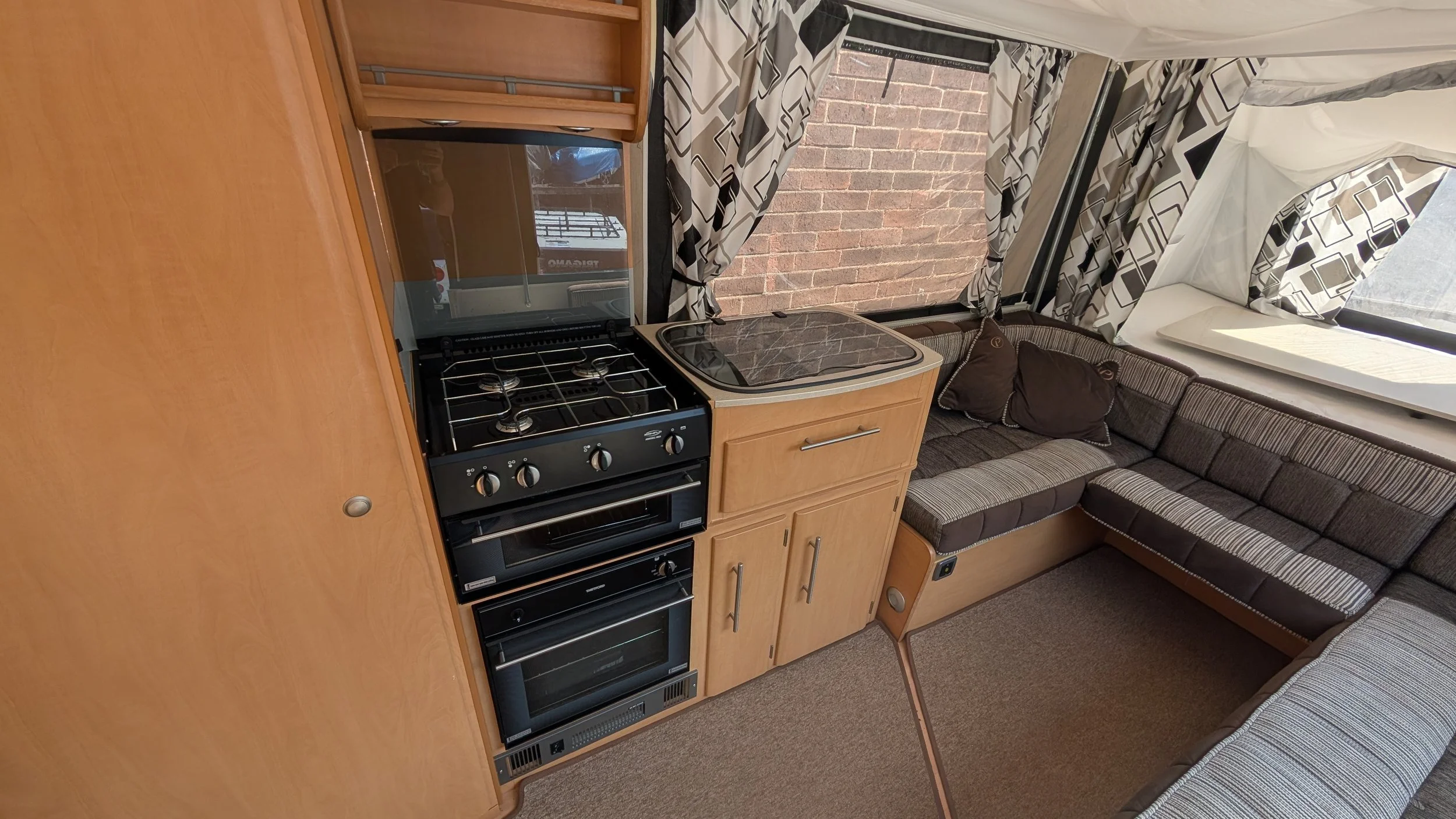 Interior of a compact living space, showing a kitchenette with a stove, oven, and microwave, a couch with pillows, and a window with black and white patterned curtains.