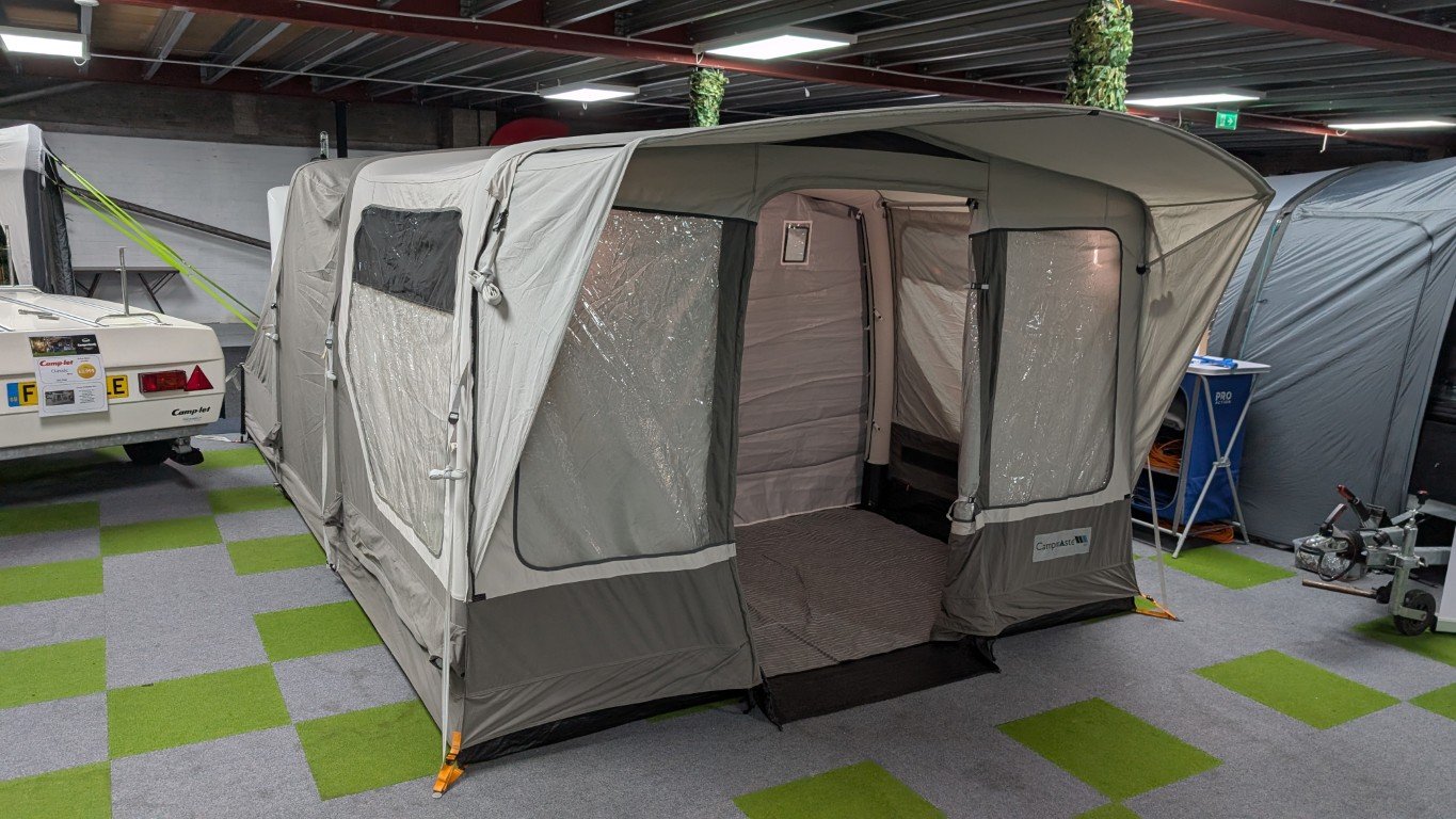 A portable camping tent with an entrance and transparent windows, set up indoors among other camping equipment and trailers, on a gray and green checkered floor.