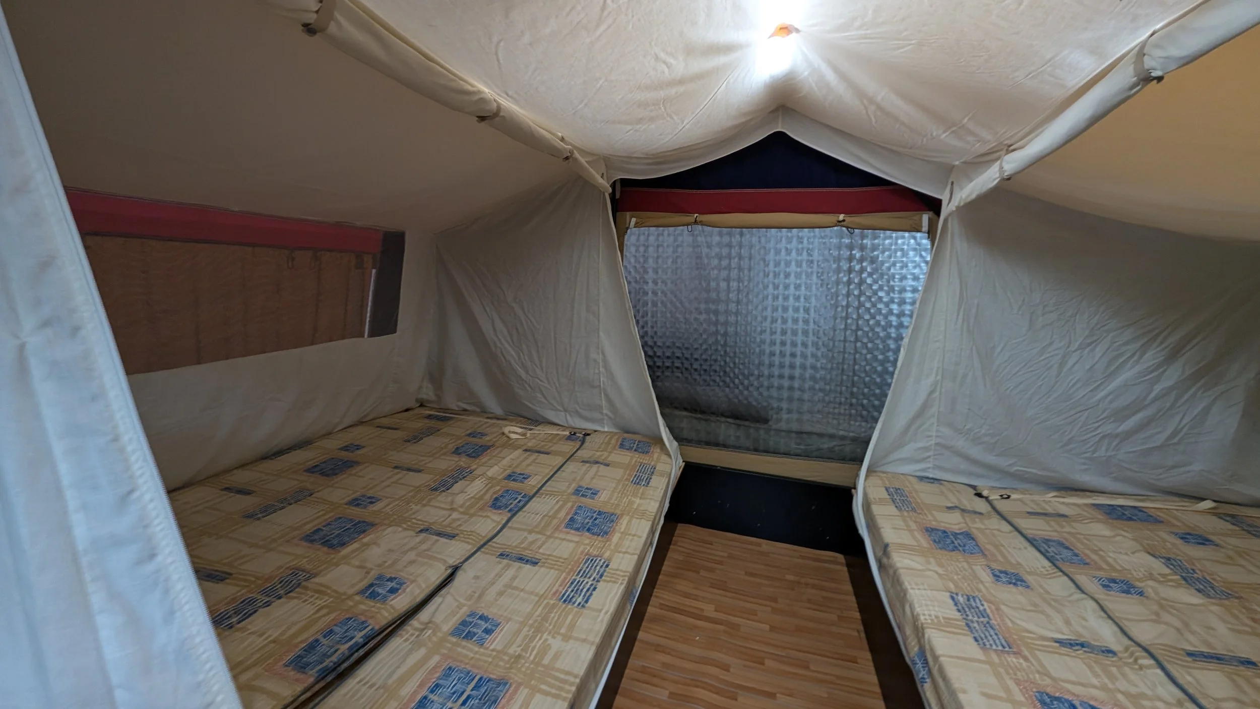 Inside a small tent with two beds, a window covered with a textured plastic screen, light-colored fabric walls, and wooden flooring.