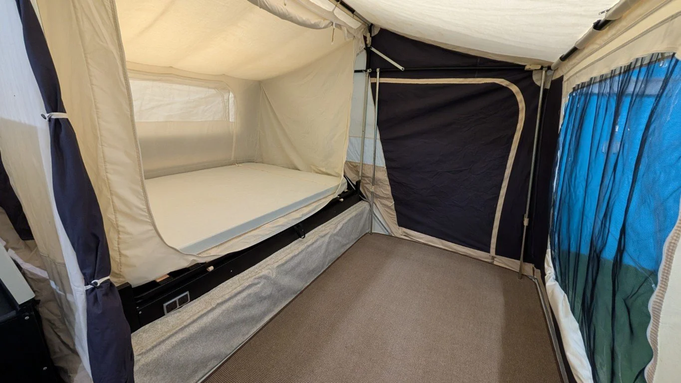 Inside of a camping tent with a foldable bed, gray flooring, and mesh windows with blue and black curtains.