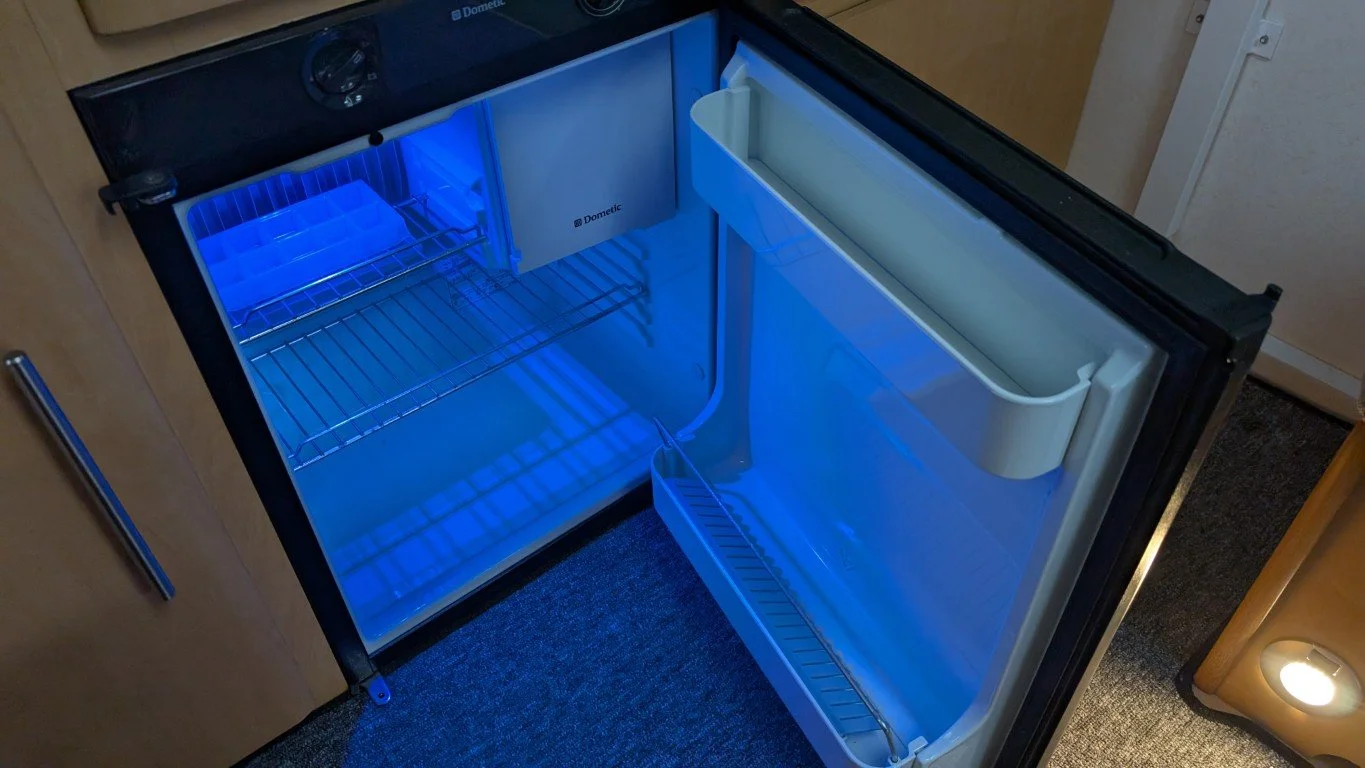 Open empty refrigerator with blue interior light, white shelves, and a wire rack at the bottom, located in a kitchen with wooden cabinets.