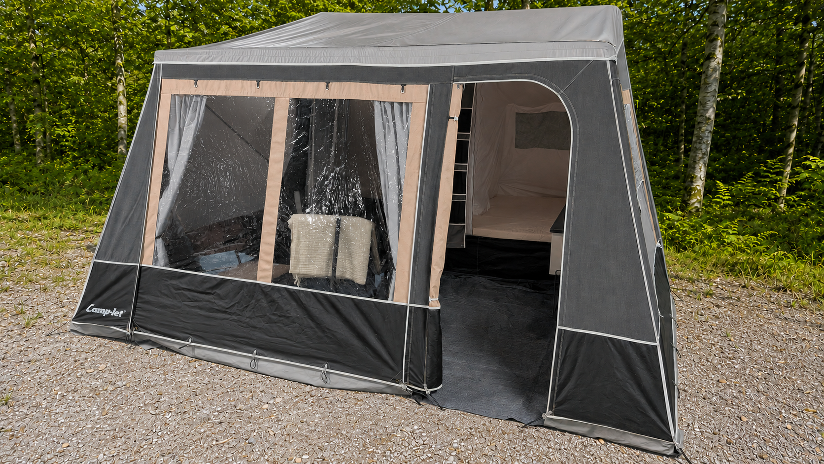 A camping tent set up outdoors on a gravel surface, surrounded by green trees.
