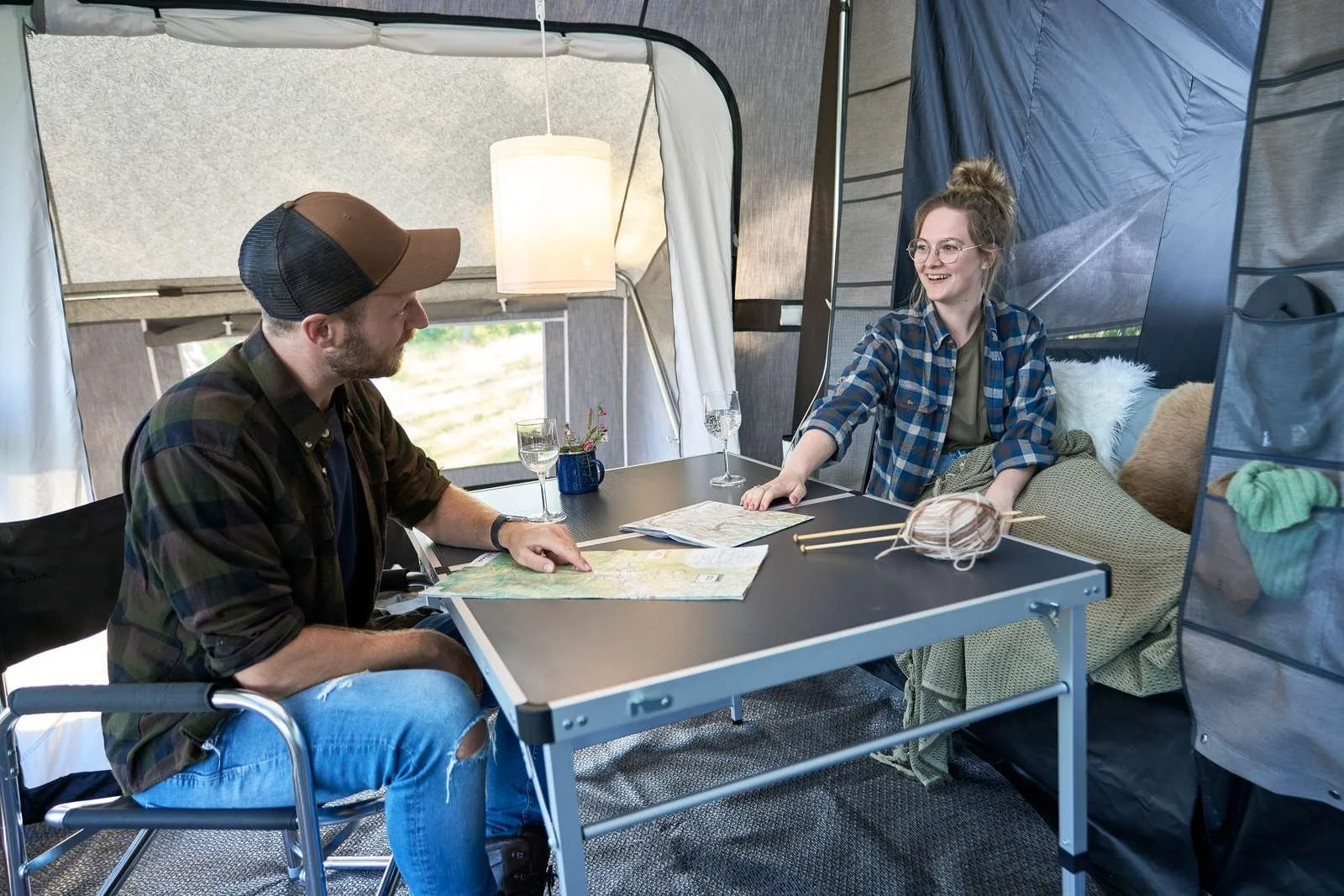 Two people sitting inside a tent at a table, engaging in conversation. The man on the left is wearing a cap and ripped jeans, and the woman on the right is smiling, wearing glasses and a blue plaid shirt. The table has maps, glasses, and yarn with kn