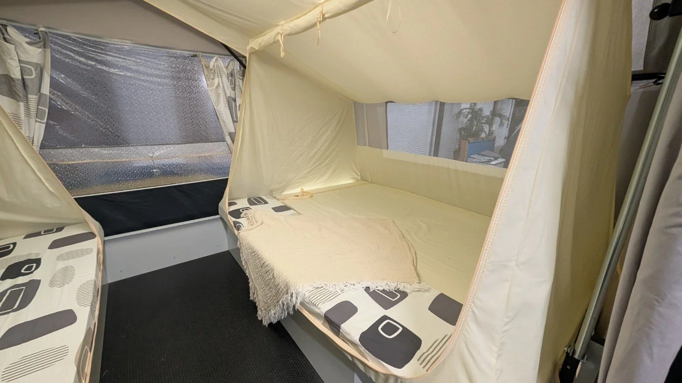 A bed inside a fabric tent with mesh windows, a blanket, and a patterned sheet, set up indoors with a view of blinds and plants outside the window.