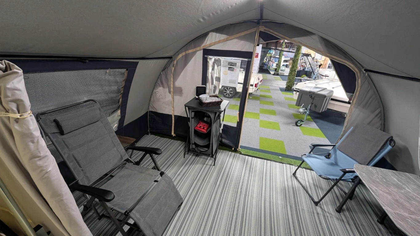 The interior of a camper or tent with three portable chairs, a small table, and a view of a campground with grass, trees, and parked vehicles.