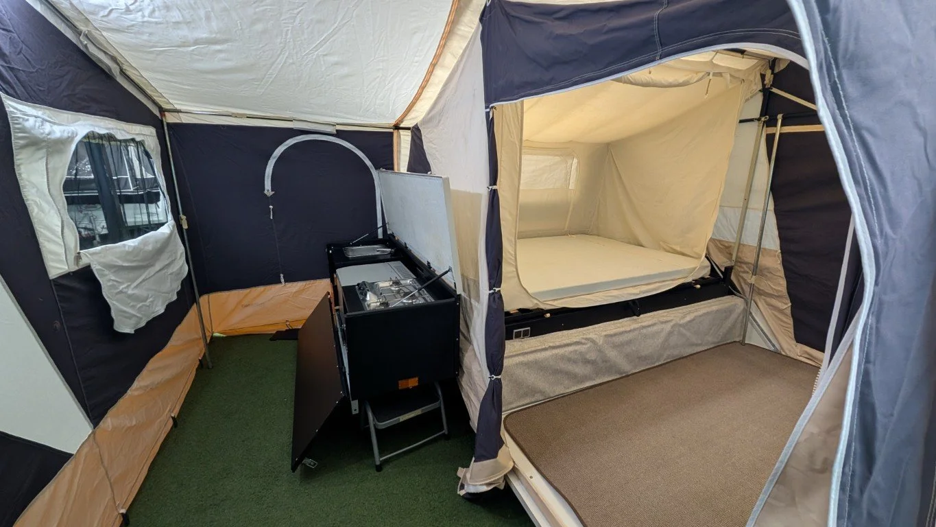 Interior of a pop-up camping tent with two sleeping areas, one with a mattress and the other with a sleeping bag, a small camping stove, and green carpet flooring.