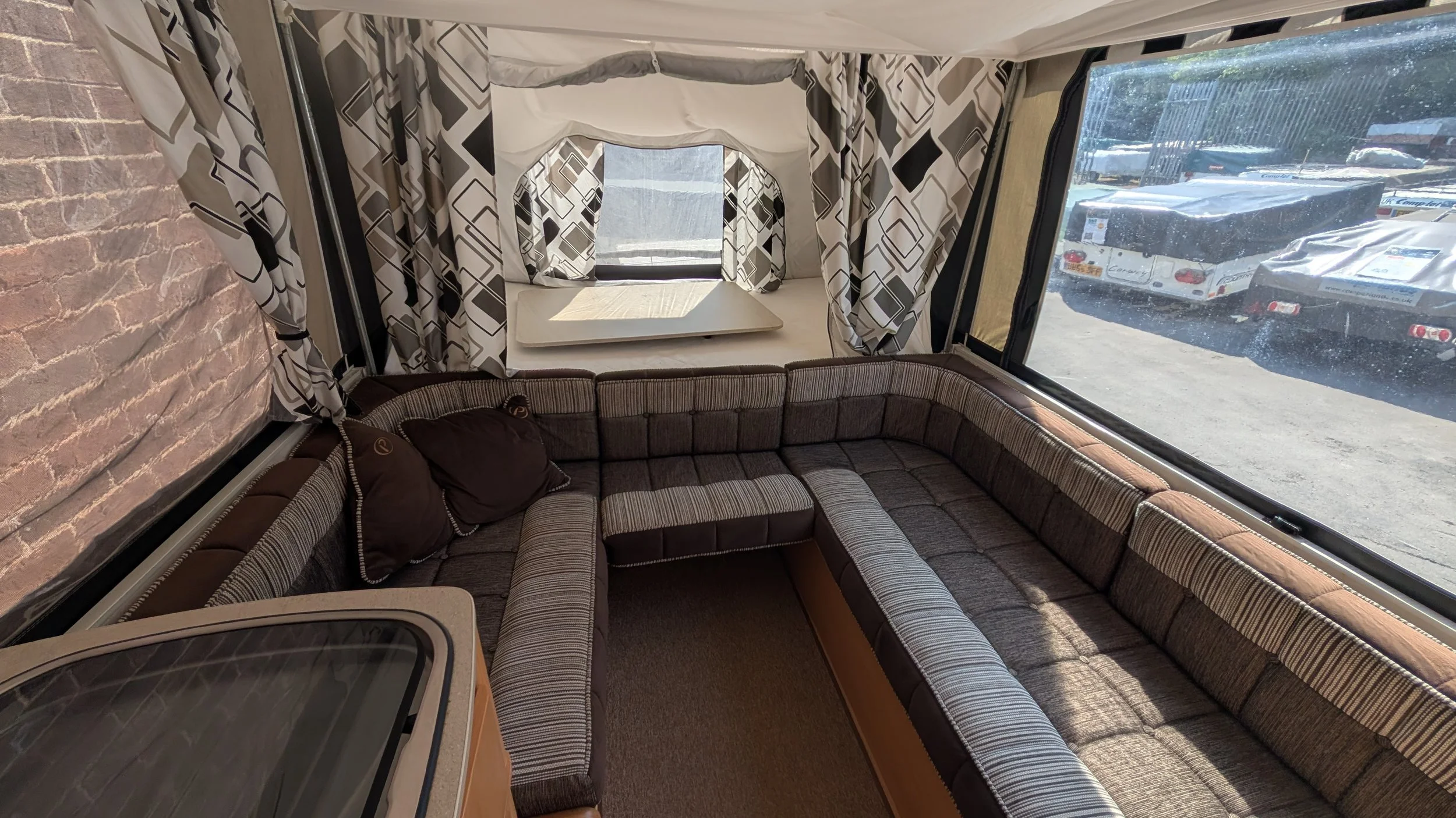 Interior of a camper trailer with a brown patterned L-shaped sofa, a window with black and white geometric curtains, and a small table in front of the window.