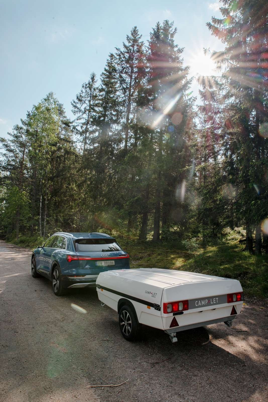 A teal-colored Audi car toing a Camp-let parked on a dirt road in a forest with tall trees and sunlight shining through.
