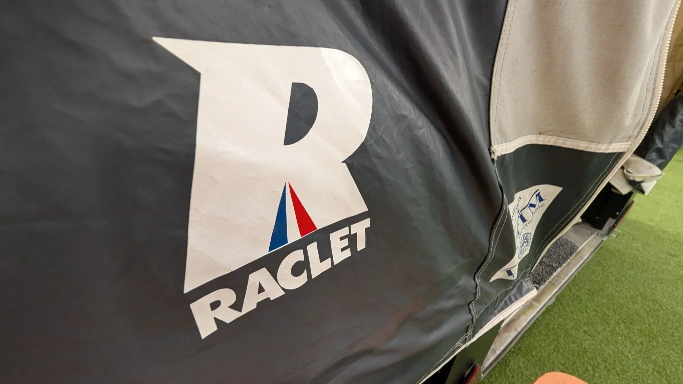 Close-up of a black fabric with a Racer Group logo featuring a stylized sailboat and the words "RACLET" beneath it. Part of a zipper and green grass are visible.