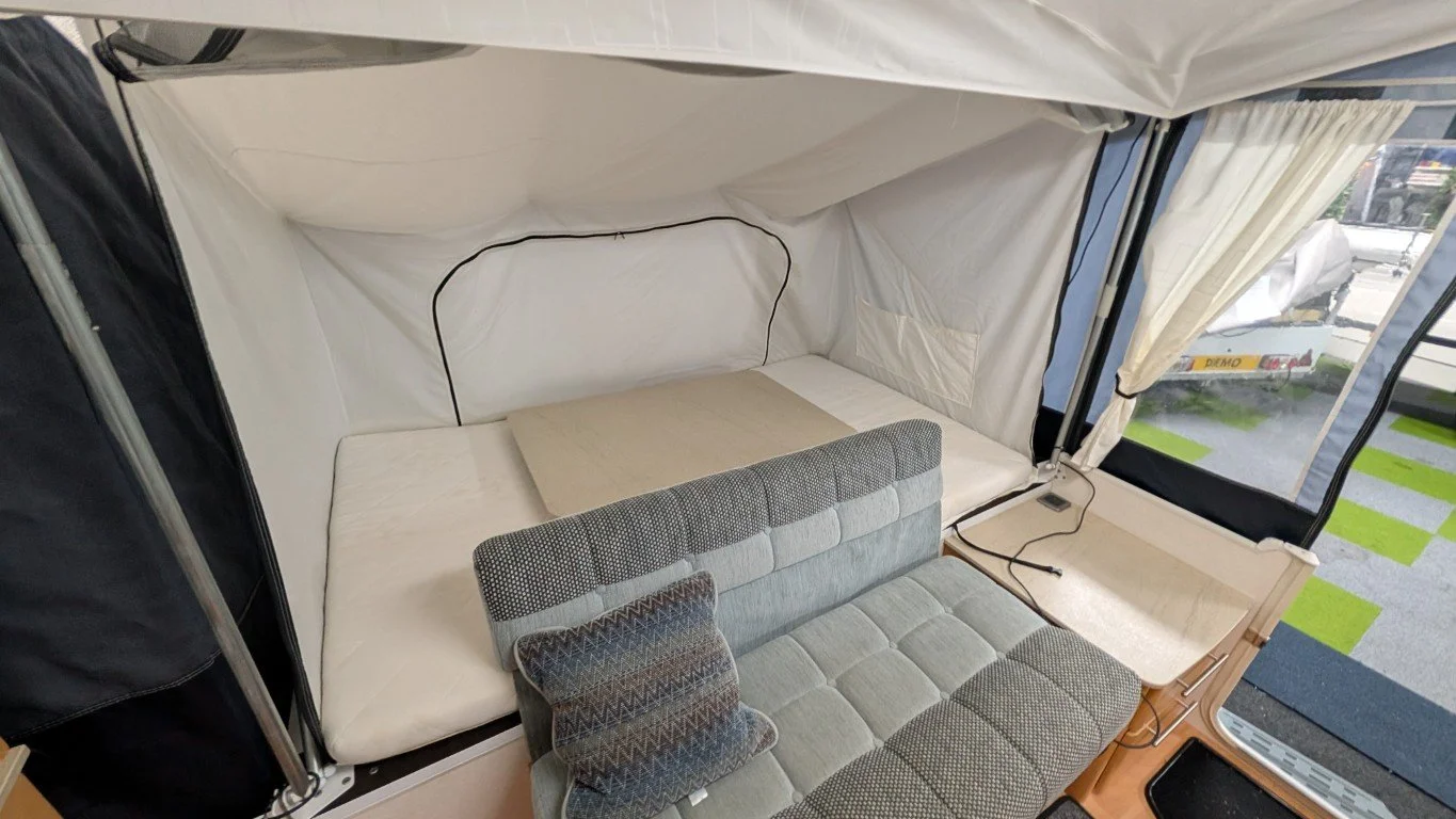 Interior of a small living space with a bed, gray patterned sofa, and large windows with curtains.