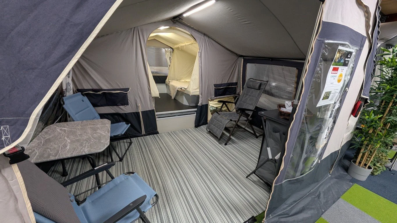 Inside of a camping tent with chairs, a small table, and a view into a sleeping area with a bed and a window.